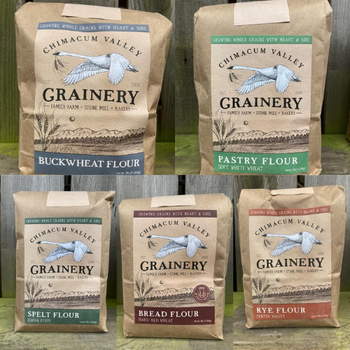 Five bags of flour from Chimacum Valley Grainery are displayed. The varieties are Buckwheat, Pastry, Spelt, Bread, and Rye, each in a rustic package.