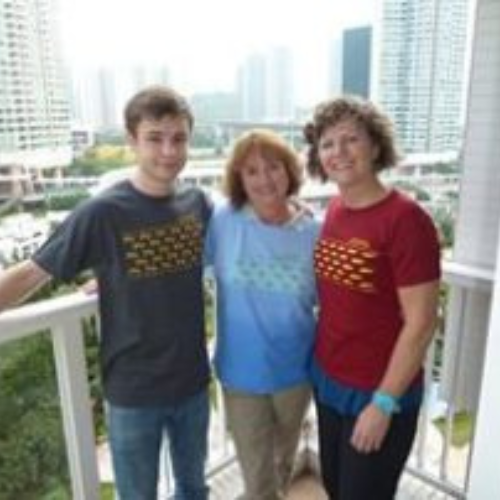 A group of three people, two women and a young man, smiling on a balcony overlooking a cityscape. They wear matching shirts, conveying a joyful and relaxed mood.