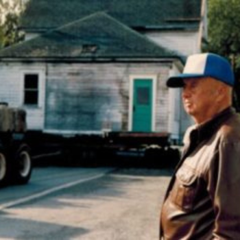 An older man in a leather jacket and blue cap stands in front of a weathered white house with a turquoise door. A truck is visible on the left.