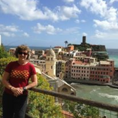 Woman smiling in a red shirt stands by a railing overlooking a picturesque seaside town with colorful buildings and a blue sky with fluffy clouds.