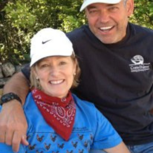 Smiling woman in a blue shirt and red bandana sits beside a man in a black shirt and cap. They're outdoors, conveying a joyful, relaxed mood.