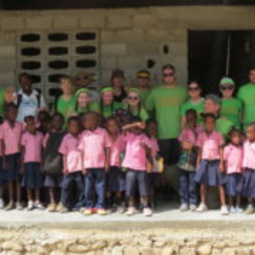 A group of smiling children in pink uniforms and adults in green shirts stand in front of a stone building, conveying joy and community spirit.
