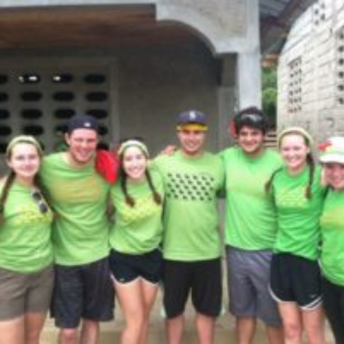 A group of seven smiling young adults wearing matching green shirts and colorful headbands poses closely together outdoors, conveying a sense of camaraderie.