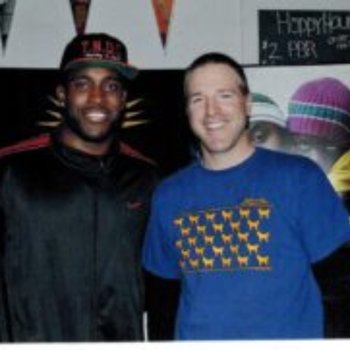 Two men are smiling, standing side by side. The man on the left wears a black cap and jacket, while the man on the right wears a blue T-shirt with yellow dogs.