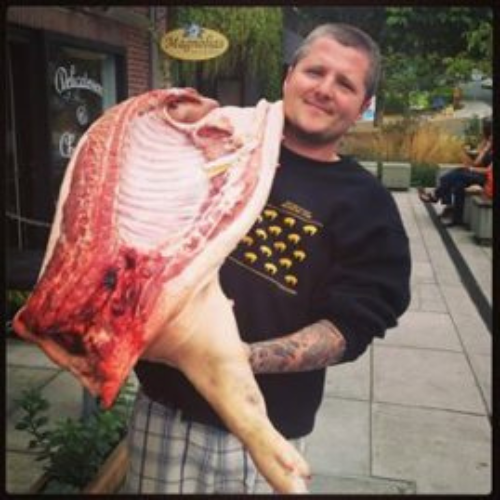 A person smiling while holding a large cut of raw meat outdoors. They're wearing a black sweatshirt and plaid shorts. The mood is lighthearted.