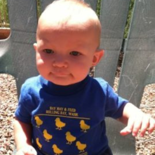 A baby sitting outdoors on a plastic chair, wearing a blue shirt with yellow chicks and text. The baby looks thoughtful under sunlight. Pebbled ground visible.
