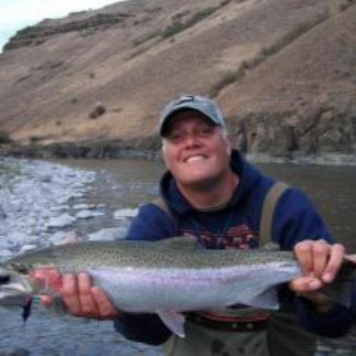 Man proudly holds a large fish, standing by a rocky river with arid hills in the background. The scene conveys satisfaction and outdoor adventure.