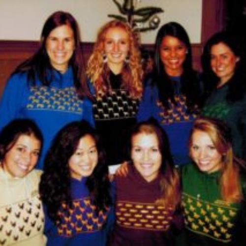 A group of eight smiling women pose together indoors, all wearing colorful sweaters with animal patterns. The atmosphere is cheerful and friendly.