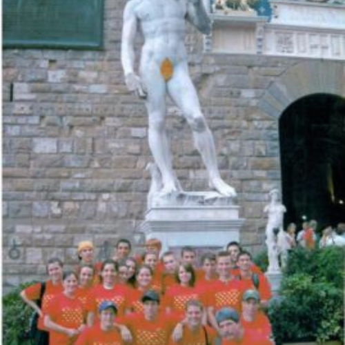A group of people in matching red shirts stands smiling near a large statue of a nude male figure. The scene looks lively and touristy, set outdoors.