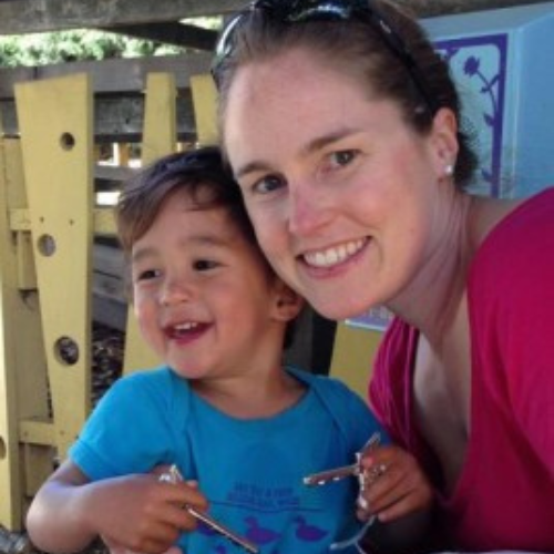 A smiling woman and child sit together outdoors. The child, wearing a blue shirt, holds metal objects. The scene feels joyful and warm.