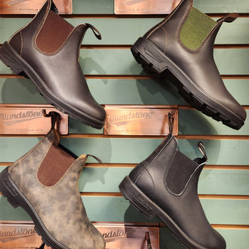 A display of four different styles of leather boots with elastic sides in brown, black, distressed, and green. Mounted on a wall with "Blundstone" plaques.