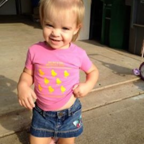 A toddler with blonde hair smiles while wearing a pink shirt with yellow ducklings and a denim skirt. She's standing on a sunlit concrete path.