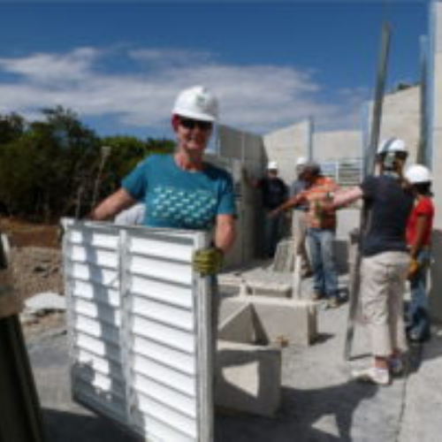 A group of people wearing hard hats work on a construction site. One person in the foreground smiles while lifting a large metal frame, conveying teamwork.