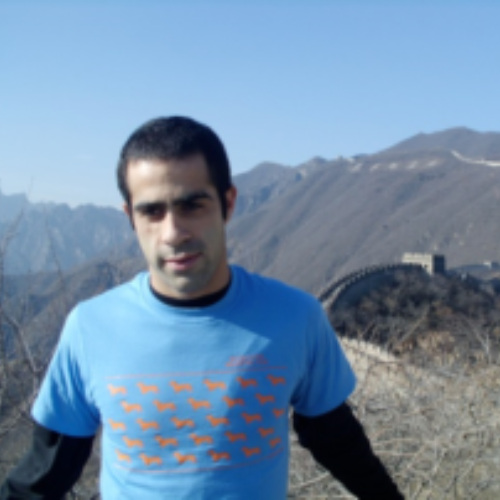 A person in a blue shirt stands in front of the Great Wall of China, with mountains in the background under a clear blue sky, conveying a sense of adventure.