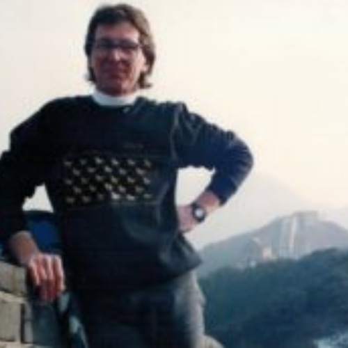 A person stands casually against a stone wall wearing a dark sweater. The Great Wall of China is visible in the misty background, evoking a sense of adventure.