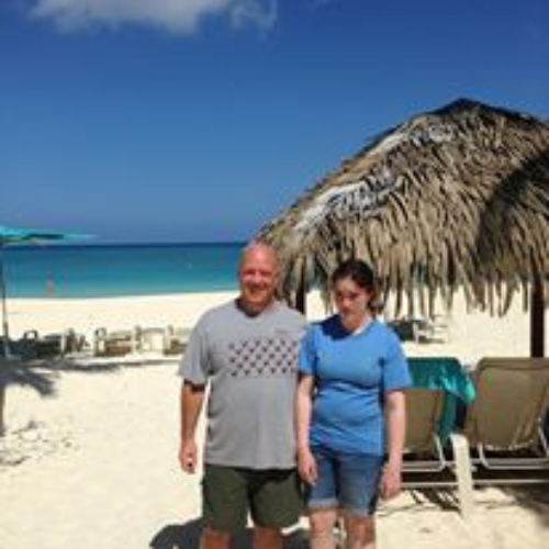 A man and woman stand on a sunny beach near a thatched umbrella, with clear blue sky and ocean in the background, conveying a relaxed vacation vibe.