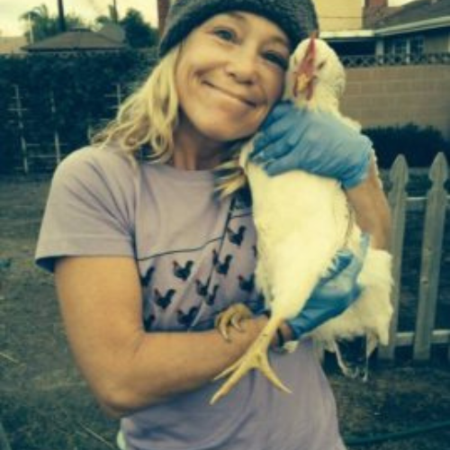 A woman in a beanie and gloves smiles warmly while holding a white chicken in a yard. The scene conveys warmth and affection.