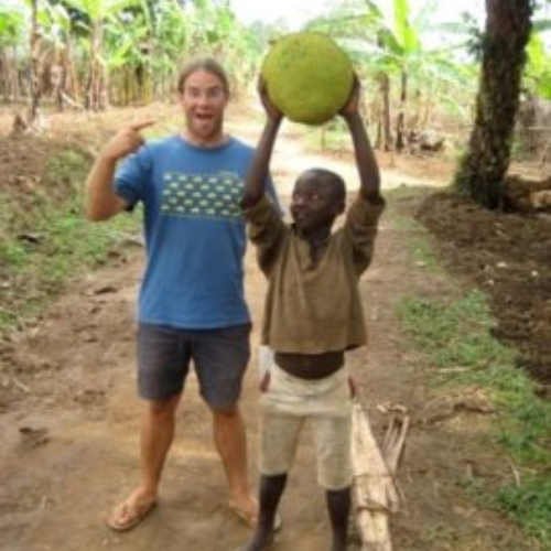Man in blue shirt excitedly points at smiling boy holding large fruit overhead. They stand on a dirt path surrounded by lush banana trees.