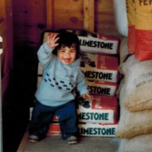 A smiling child in a light blue sweater waves while sitting on stacked limestone bags in a wooden shed. The scene feels playful and cheerful.