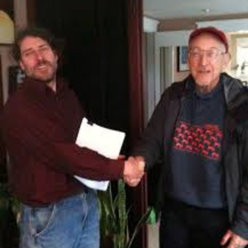 Two men are shaking hands indoors, smiling warmly. One holds a document, wearing a dark shirt and jeans; the other wears a cap and dark jacket, creating a friendly, positive atmosphere.