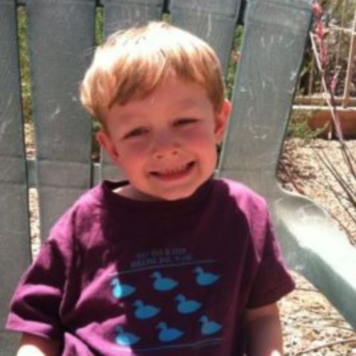 Young boy with blond hair smiling while sitting on a light green chair. He wears a purple shirt with blue ducks. Sunny outdoor background.