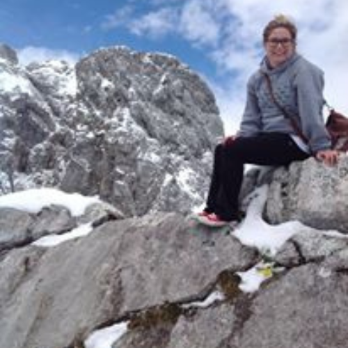 A person in winter clothing sits on a snowy rock, smiling with a towering, snow-covered mountain in the background under a clear blue sky.
