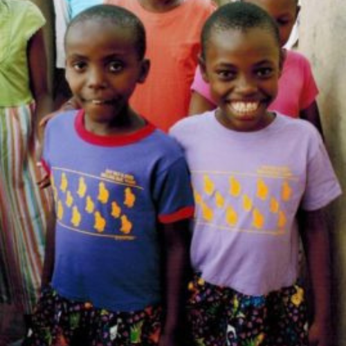 Two smiling children stand side by side, wearing colorful skirts and shirts with yellow prints. Other children and bright colors are in the background.