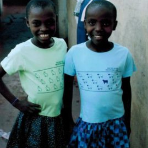 Two young girls stand close together, smiling broadly, wearing patterned skirts and light-colored T-shirts. The background is a simple outdoor setting.