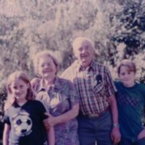 A family photo of two children and two elderly adults standing outdoors. The children wear casual clothes; the adults are smiling, amidst a sunny garden backdrop.