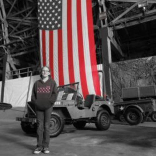 A person stands in front of a military jeep inside an industrial building, with a large American flag hanging behind. The image conveys a sense of history and patriotism.