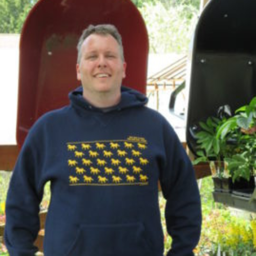 A smiling man in a navy hoodie with yellow horse patterns stands outdoors. Behind him, red and black wheelbarrows and greenery are visible, suggesting a garden setting.