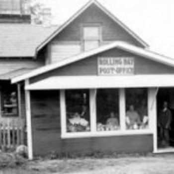 Black and white photo of an old post office with a sign reading "Roehl's Bay Post Office." Three people visible in the window, conveying a historic, nostalgic atmosphere.