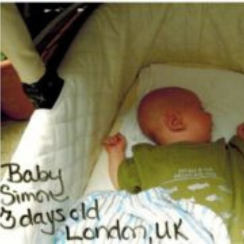 A newborn baby lies asleep in a stroller, wearing an olive green outfit. Handwritten text reads 