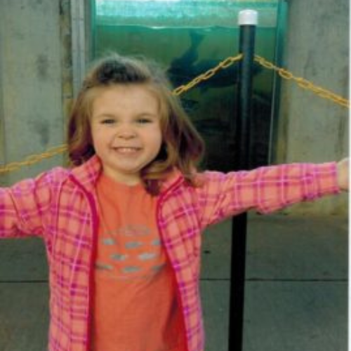 A smiling child in a pink plaid jacket stands with arms outstretched in front of an aquarium, with a shark visible in the tank behind them.