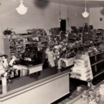 Vintage black-and-white photo of a quaint grocery store interior with shelves of canned goods, a wooden counter, a cash register, and floral displays.