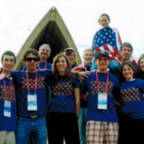 A group of cheerful people wearing matching blue shirts with printed designs pose in front of a triangular structure. One person is draped in a flag.