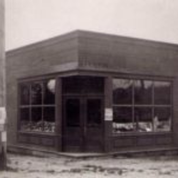 A vintage black-and-white photo of a corner store with large windows displaying goods. The wooden facade and signage suggest an early 20th-century setting.