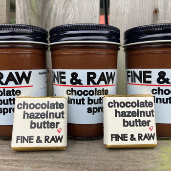 Three jars of Fine & Raw chocolate hazelnut butter spread are lined up against a wooden backdrop. In front, two chocolate bars with matching labels.
