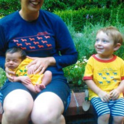 A person in a navy shirt holds a sleeping baby in a yellow outfit. Beside them, a smiling toddler in a matching shirt and striped pants sits on a ledge.