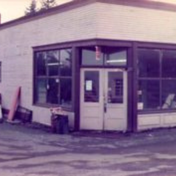 A vintage corner store with large windows and a double door, showcasing a weathered exterior. The scene evokes a nostalgic, rustic feel.