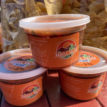 Three tubs of Ardell Salsa labeled with red-orange packaging are stacked on a wooden table, surrounded by bags of tortilla chips, conveying a fresh, appetizing feel.
