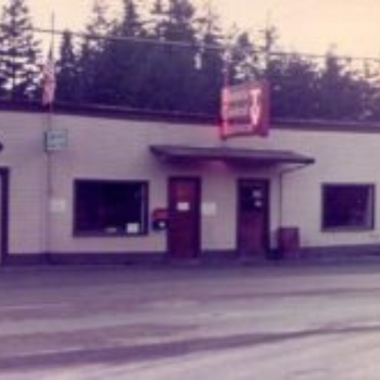 Vintage storefront with red signage and American flag, set against a backdrop of tall trees under a soft, pinkish sky, evoking nostalgia.