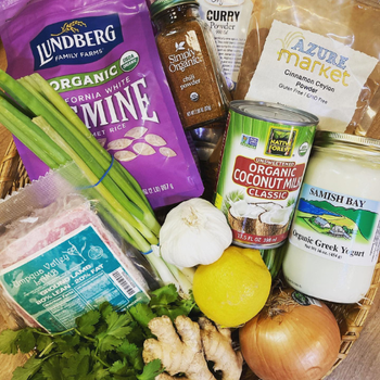A collection of organic ingredients in a basket, including jasmine rice, curry and cinnamon powders, coconut milk, Greek yogurt, garlic, lemon, green onions, cilantro, fresh ginger, and an onion.