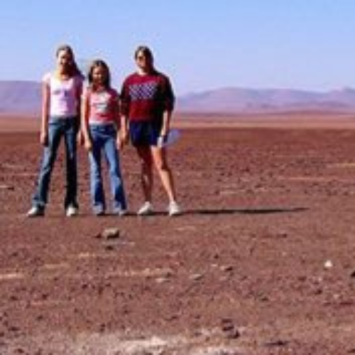 Three people stand in a vast, barren desert landscape under a clear blue sky. The distant mountains in the background create a sense of isolation.