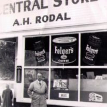 A man in a coat stands smiling outside the "Central Store," with Folger's coffee ads displayed on the windows. The scene conveys a sense of community and nostalgia.