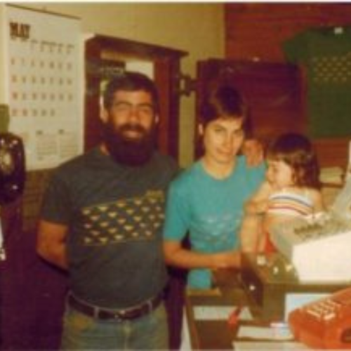 A vintage photo shows two adults and a child in a warmly lit room. The man, with a beard, stands smiling; the woman is holding the child. A calendar and rotary phone are visible, evoking a nostalgic atmosphere.