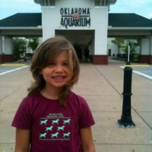 Young child with long hair smiling in front of Oklahoma Aquarium entrance, wearing a maroon shirt. The sky is overcast, creating a subdued atmosphere.