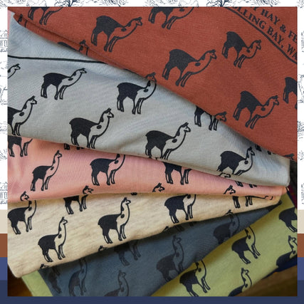 Bay Hay ShirtsFolded fabrics in shades of red, blue, pink, beige, and green, each adorned with a repeating llama pattern, evoking a playful and cozy vibe.