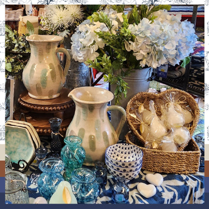 Gifts & ToysCeramic pitchers and blue glass vases on a table with a floral tablecloth are surrounded by white hydrangeas and a basket of wrapped soaps, creating a cozy and elegant display.