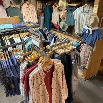 Clothing store interior with colorful shirts, hats, and patterned fabrics neatly arranged on racks. The atmosphere is casual and welcoming.